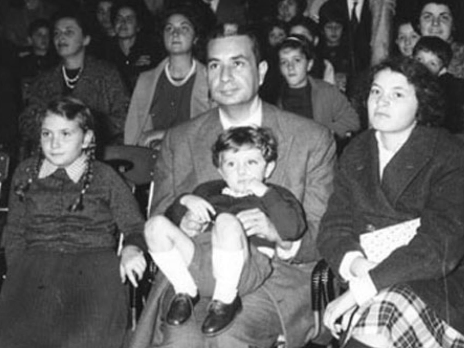 Aldo Moro com seus filhos durante um evento público, em um raro momento de descontração familiar. Moro e Eleonora tiveram quatro filhos: Maria Fida, Agnese, Anna e Giovanni. Esta fotografia mostra Moro não como o arquiteto do Compromisso Histórico ou o líder da Democracia Cristã, mas como um pai amoroso, segurando uma de suas crianças no colo enquanto outra se senta ao seu lado. Para seus filhos, Aldo Moro não era um estadista: era simplesmente "papai", o homem que voltava para casa após longas sessões no Parlamento, que ajudava com as lições de casa, que acreditava que a família era o refúgio sagrado contra as tempestades da política. Durante os 55 dias de sequestro, as crianças de Moro viveram um pesadelo. Elas assistiram, impotentes, enquanto sua mãe implorava ao governo que salvasse o pai.