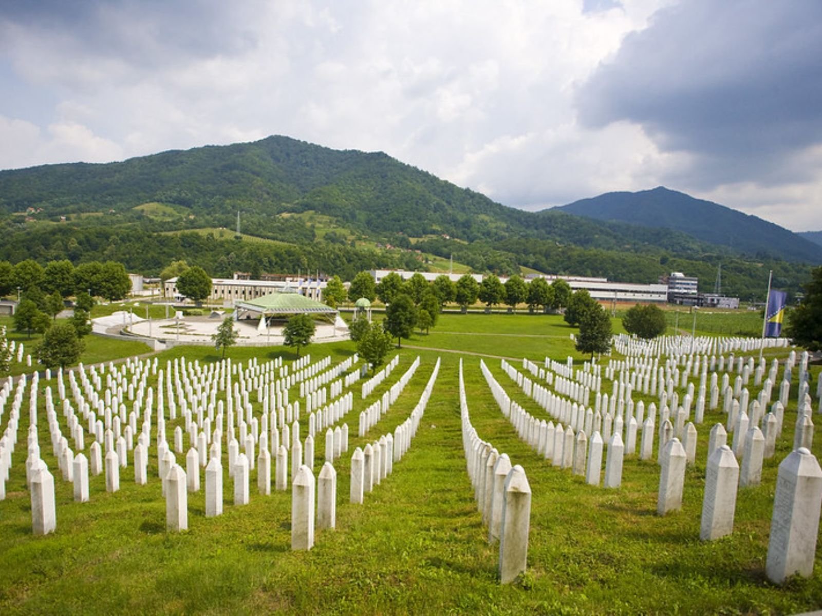 O Memorial e Cemitério de Srebrenica-Potočari, onde mais de 6.600 vítimas identificadas do genocídio de julho de 1995 foram enterradas. Esta vista panorâmica captura a magnitude visual do horror: milhares de lápides brancas de mármore se estendem em fileiras perfeitamente alinhadas sobre o gramado verde, contrastando com as montanhas verdejantes dos Bálcãs ao fundo.
