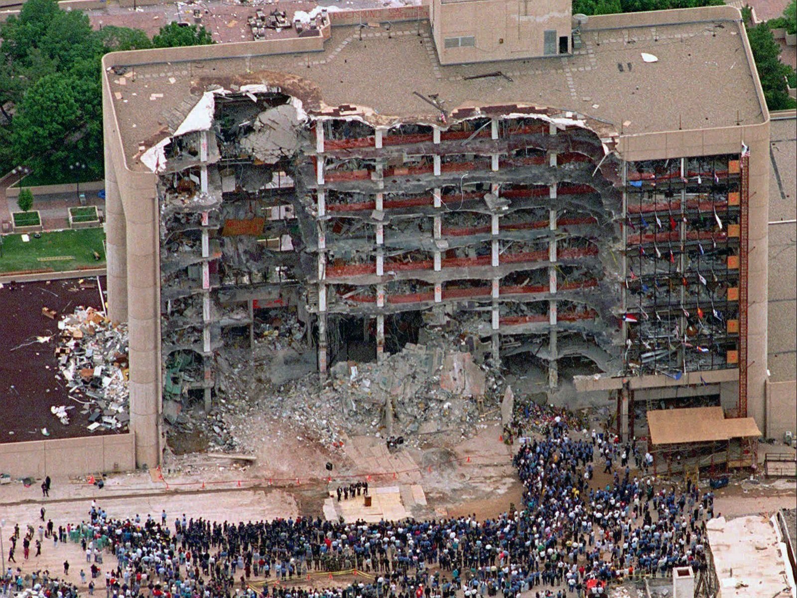 A fachada norte do edifício federal Alfred P. Murrah em Oklahoma City, completamente destruída após a explosão do caminhão-bomba em 19 de abril de 1995. A imagem revela a escala da devastação causada pela bomba de Timothy McVeigh, que arrancou nove andares do prédio onde funcionavam escritórios federais e uma creche com 19 crianças. Centenas de pessoas se aglomeram nos escombros em operações de resgate que duraram semanas. Fonte: Britannica.