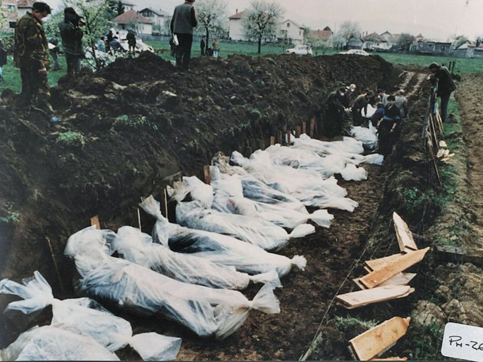 Enterro em massa de vítimas da Guerra da Bósnia em uma vala coletiva. Dezenas de corpos envoltos em sacos brancos e plásticos são colocados lado a lado em uma trincheira cavada em campo aberto, enquanto investigadores forenses e trabalhadores documentam e supervisionam o processo. O código "PR-261" visível no canto inferior direito sugere que esta é uma fotografia de arquivo forense, provavelmente parte de investigações de crimes de guerra.