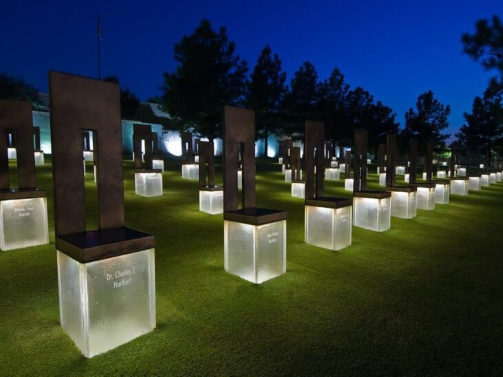 O Memorial Nacional de Oklahoma City, onde 168 cadeiras vazias de bronze e pedra representam cada vida perdida no atentado de 19 de abril de 1995. As 19 cadeiras menores simbolizam as crianças mortas, dispostas em nove fileiras correspondentes aos andares do prédio destruído. Cada cadeira carrega o nome de uma vítima gravado na base, criando um santuário silencioso que serve como lembrete perpétuo do custo humano do ódio extremista e da violência política normalizada. Fonte: The Journal Record.