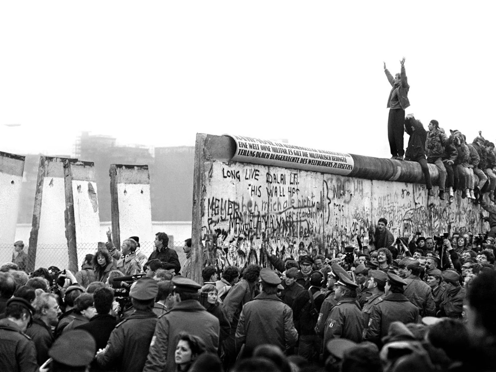 10 de novembro de 1989, dia seguinte à abertura dos portões. Centenas de berlinenses ocupam o topo do Muro de Berlim em um ato simbólico de desafio e celebração. Uma pessoa ergue os braços em gesto de vitória no ponto mais alto, enquanto multidões se aglomeram tanto sobre quanto ao redor da barreira que os havia dividido por 28 anos. O Muro, outrora símbolo intocável de opressão — onde qualquer pessoa que se aproximasse demais arriscava ser baleada —, agora se transformou em palco de uma festa popular espontânea.