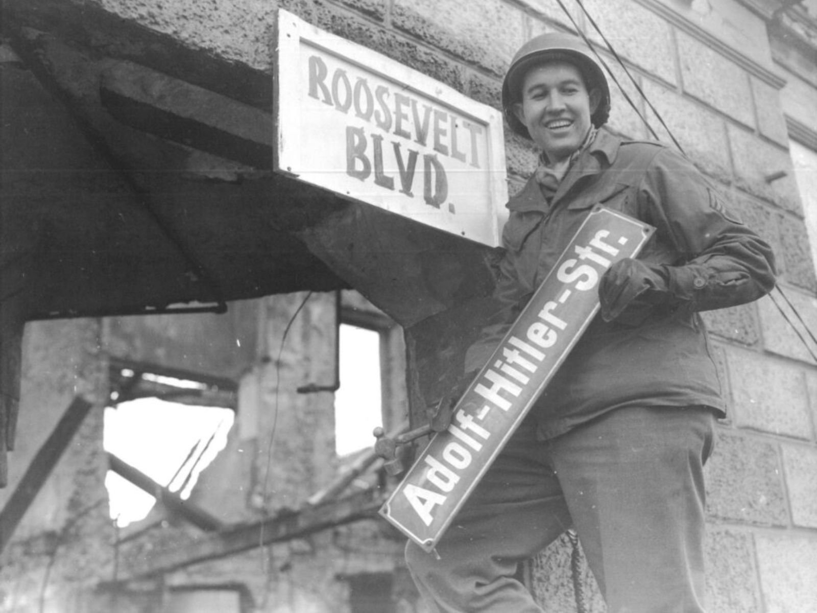 Um soldado americano substitui uma placa da "Adolf-Hitler-Str." por "Roosevelt Blvd.", um ato simbólico da desnazificação e da imposição da cultura americana.