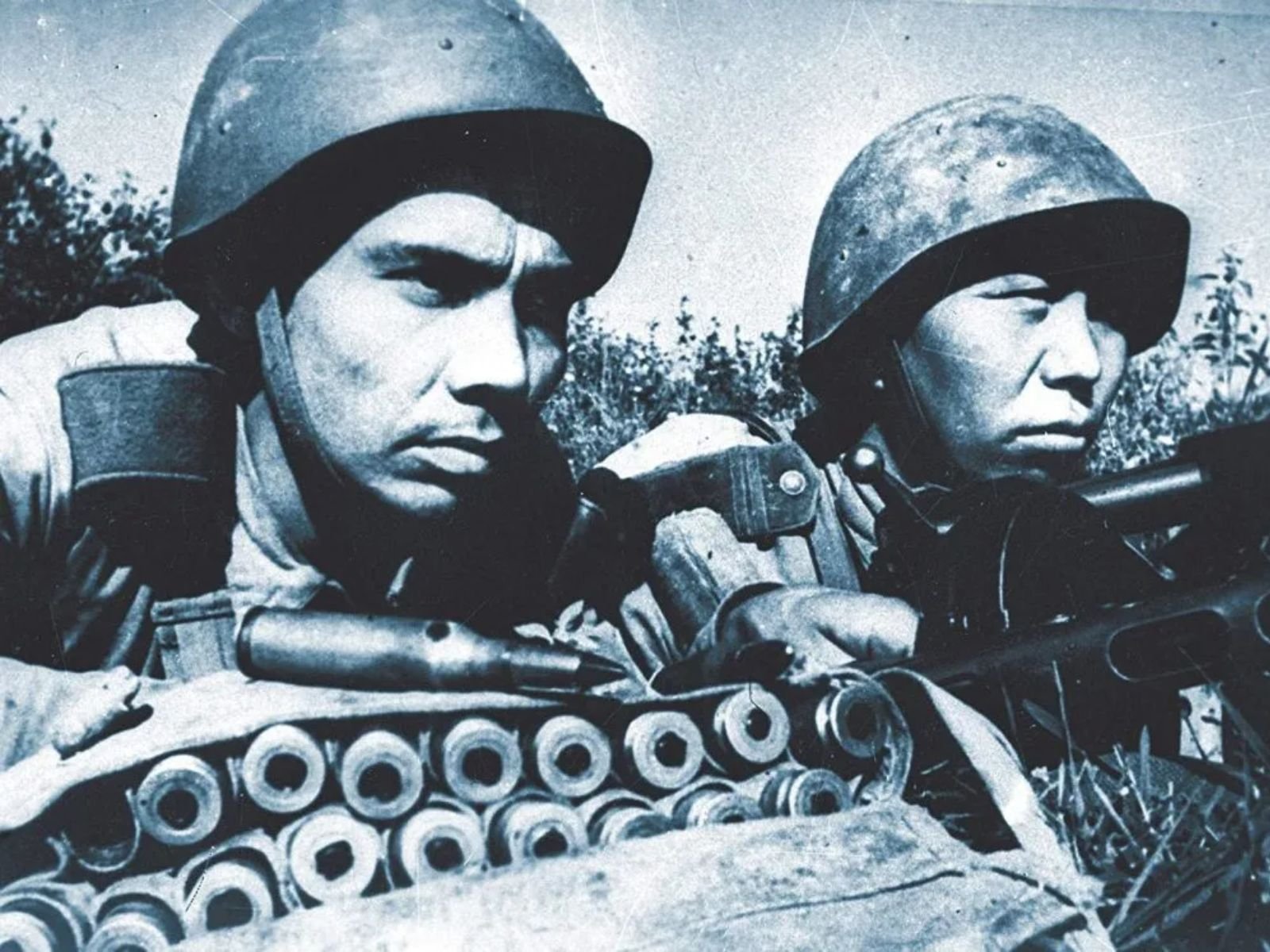 Fotografia em formato paisagem mostrando dois soldados do Exército Vermelho de origem centro-asiática (provavelmente cazaques ou uzbeques) em posição de combate. Os soldados usam capacetes soviéticos padrão e estão equipados com rifles e cinturões de munição. A imagem tem uma tonalidade azulada característica de fotos coloridas artificialmente da época.