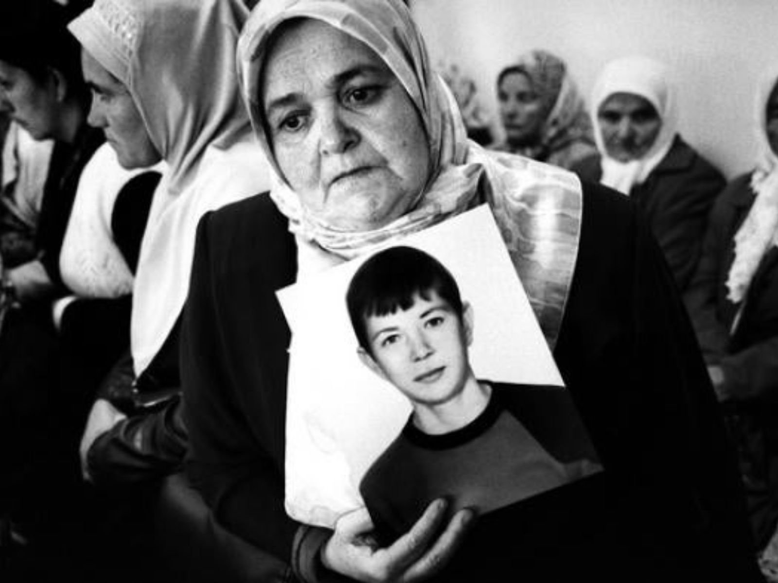 Uma mulher bósnia muçulmana segura a fotografia de seu filho desaparecido, um dos milhares de homens e meninos mortos no genocídio de Srebrenica em julho de 1995. A imagem, em preto e branco, captura a dor e a busca por justiça das "Mães de Srebrenica", uma associação de mulheres que perderam seus entes queridos no massacre.