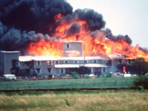Cerco de Waco: David Koresh, Terror e Tragédia O complexo dos Ramo Davidianos em Monte Carmelo consumido pelas chamas em 19 de abril de 1993, após 51 dias de cerco federal. Esta imagem icônica captura o momento final da tragédia que matou 84 pessoas, incluindo David Koresh e 25 crianças. O incêndio começou após agentes federais lançarem gás lacrimogêneo e romperem as paredes com tanques militares. A controvérsia sobre quem iniciou o fogo - se os próprios davidianos ou as ações federais - persiste até hoje.