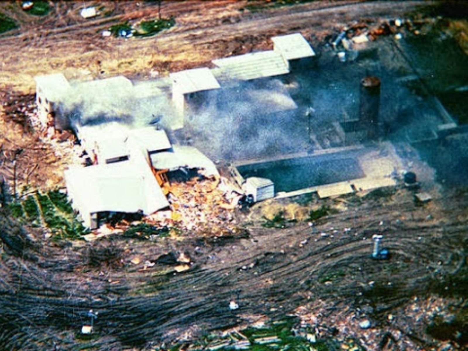 Vista aérea devastadora do complexo dos Ramo Davidianos após o incêndio de 19 de abril de 1993. A imagem revela a extensão completa da destruição em Monte Carmelo, onde David Koresh liderava sua comunidade religiosa desde o final dos anos 1980. O que antes era o "Rancho Apocalipse" - como Koresh havia renomeado a propriedade - tornou-se um símbolo das tensões entre liberdade religiosa e aplicação da lei nos Estados Unidos.